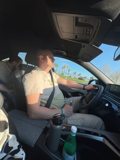 The image depicts the interior of a vehicle on a clear, sunny day in Inca, Spain. A male driver, with short hair, wears a light-colored short-sleeved t-shirt, tan cargo pants, a dark wristband on his left wrist, and multiple beaded bracelets on his right. He is secured by a black seatbelt, holding the black steering wheel with his right hand, and a dark pen-like object in his left hand, looking forward and slightly right. Sunlight illuminates his left side. A male rear passenger, bearded and wearing a light-colored jacket, is partially visible behind the driver. The vehicle's interior features a black headliner, overhead console, and a grey fabric-covered center console. Two beverages are in the front cup holders: a dark grey insulated water bottle and a green San Pellegrino sparkling water bottle. Outside the vehicle, a bright blue sky is visible above a grassy embankment densely populated with tall palm trees, and some leafless deciduous trees are in the distant background.
