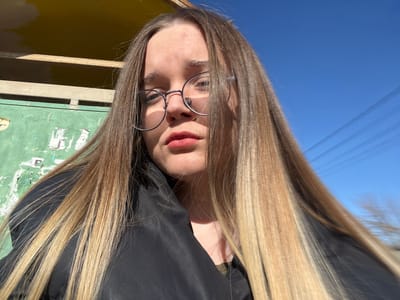 A close-up, low-angle self-portrait features a young female subject with light-colored, long, straight hair, styled with an ombre effect transitioning from brown at the roots to blonde at the ends. She wears round, silver-rimmed eyeglasses and a black collared jacket over what appears to be a dark green shirt. Her lips are covered with a red lipstick, and a faint mark is visible on her forehead. She maintains direct eye contact with the camera. The background on the left consists of a weathered, pale green wall with peeling paint and remnants of white paper affixed to it, topped by a section of a yellow structure. To the right and top, a clear, bright blue sky is visible, traversed by several thin, dark horizontal lines, consistent with overhead wires. The scene is illuminated by direct sunlight, casting shadows on the subject's face. The image depicts an outdoor urban setting in the city of Большевик, Russia.