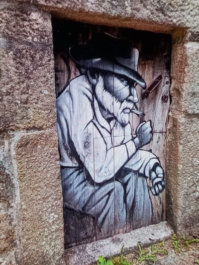 A monochromatic mural, rendered in shades of grey and black, is depicted on a recessed wooden panel set within a rough-hewn stone wall in Aktash, Uzbekistan. The central subject is an older male figure, seated, with a prominent beard, mustache, and a wide-brimmed hat. He is shown holding a pipe in his left hand, and a lit cigarette or cigar extends from his mouth, oriented to the right. His right arm rests on a bent knee. The wooden panel exhibits distinct vertical wood grain, and a metallic latch mechanism is present on its upper right side. The surrounding wall is constructed from irregular, light-brown textured stone blocks. A narrow, flat stone ledge forms the base of the recessed panel, below which sparse green vegetation grows on the ground.