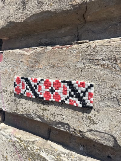 A mosaic strip made of small red, black, and white square tiles is affixed to a weathered, light-colored concrete wall. The mosaic forms a geometric pattern of alternating rows of black and red squares, interspersed with white squares. The concrete wall appears rough and cracked, with some patches of discoloration and moss. The mosaic is positioned horizontally across the visible portion of the wall. The scene suggests an urban outdoor environment, potentially a public space in Novaya Usman, Russia. The lighting indicates it is daytime.