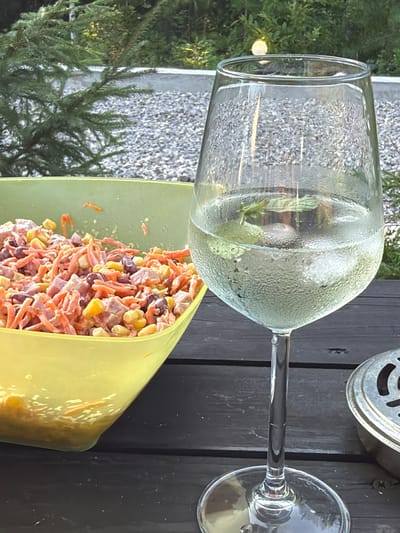 The image depicts an outdoor scene on a dark wooden table, likely in Kuliki, Russia, with a glass of white wine filled with ice and mint in the foreground. To the left of the wine glass is a bright yellow bowl overflowing with a colorful salad made of shredded carrots, corn, ham, and kidney beans, all coated in a creamy dressing. The background features lush green trees and a path covered with gray gravel. A small, metallic object, possibly an ashtray, is partially visible on the right side of the table. The overall ambiance suggests a casual outdoor gathering or meal.