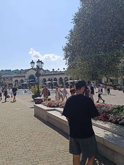 The image captures a sunny day in Serravalle Scrivia, Italy. The main subject is a man facing away from the camera, wearing a black t-shirt and gray shorts, with a black baseball cap on his head. He stands near a raised concrete planter filled with colorful flowers. In the background, a bustling public square is visible with many people strolling around. Shops and stores are aligned in the back, identifiable by their signage (e.g., "MOSCHINO," "ZEGNA," "VERSACE"). The square features a fountain, lamp posts, and a large tree that provides shade. The ground is paved with bricks. The sky is light blue with a few scattered clouds.
