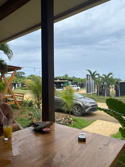 The image shows a view from a covered patio, looking out onto a dirt path leading to a street. There is a silver SUV parked on the left side of the path. The car is parked facing the camera with the driver's side visible. In the background, the path leads to several buildings, including a small shop on the right. There are palm trees and other greenery surrounding the buildings, the path, and the patio. The patio is made of wood and has a wooden table with a glass of orange juice, a purse, and a pack of cigarettes on it. There is a woman sitting at the table with a blurry upper body. The sky is overcast with clouds. The location is Las Escobas del Venado, Panama, based on the coordinates (latitude: 7.432, longitude: -80.189).