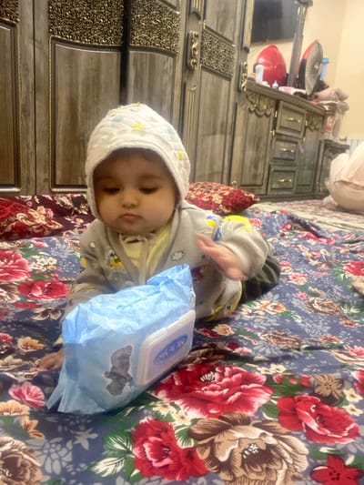 An infant, wearing a white quilted bonnet and a light-colored full-body sleepsuit with yellow accents, is positioned on a dark blue floral patterned fabric spread on the floor in Galaosiyo, Uzbekistan. The child's left hand is extended towards a light blue package of "Me to You" wet wipes, which features a gray teddy bear graphic and a white plastic dispenser. The child is looking downwards, towards the wipes. Behind the child, two patterned pillows are visible. The background features dark wood furniture with gold-colored ornate details, including a large wardrobe on the left and a dresser with drawers on the right. Items on the dresser include a red appliance, a black fan, and a white bottle. A mirror above the dresser reflects part of the ceiling. On the far right, a person's lower body, clad in light-colored attire, is partially visible, kneeling or sitting on the carpeted floor.