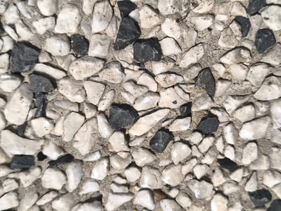 This is a close-up, overhead, high-angle, full-frame, eye-level shot of a cobblestone path in Boskoop, Netherlands. The path is composed of a mixture of light-colored, irregular-shaped stones and dark-colored, irregular-shaped stones, set in a rough, sandy-colored mortar. The stones vary in size and shape, with some being rounded and others more angular. The surface appears to be dry. The lighting suggests it is daytime. There are no people, animals, or other objects visible in the image.