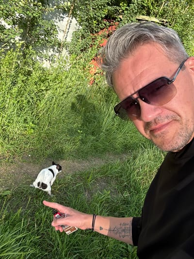 The image shows a man wearing sunglasses, with graying hair, and a goatee, looking towards the camera. He is wearing a black shirt and has a tattoo on his arm. A small black and white dog is on the grass in front of him, and the man is pointing towards it with his right hand, which is holding a pack of cigarettes. The setting appears to be outdoors, in a grassy area with some vegetation. The lighting suggests it is daytime, likely sunny, given the strong shadows. The scene is set in Лесные Поляны, Russia. The man's expression is neutral or slightly amused. The text visible on the cigarette pack is "Chapman".