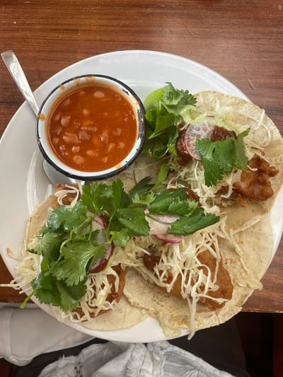 A plate of three tacos dominates the frame. Each taco is filled with fried fish, shredded cabbage, radish slices, and cilantro. A small bowl of beans in a reddish-orange sauce sits alongside the tacos, with a spoon resting near it. A lime wedge is nestled amongst the taco ingredients. The food rests on a white plate, which is placed on a wooden surface. The edge of a white cloth is visible at the upper right of the image.