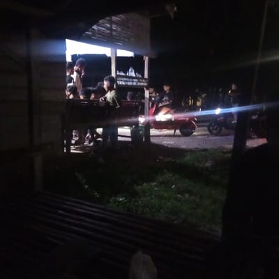 At night in Matangmeunye, Indonesia, a group of people are gathered under a wooden structure. A red scooter with the word "GODA" on it is parked nearby. There is another vehicle with its headlights on in the background. The area in front of the structure has grass and a dark corrugated metal surface.