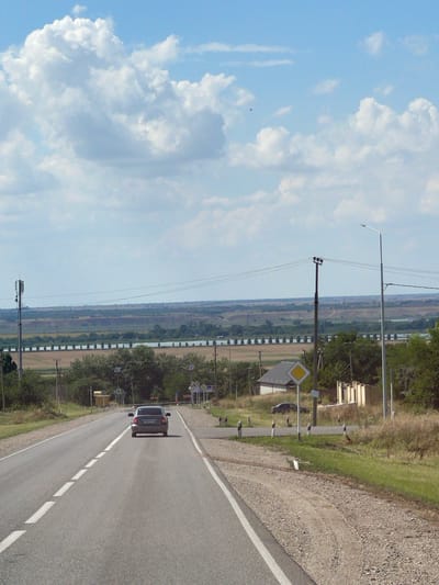 A dark-colored sedan drives away from the camera on a two-lane asphalt road in a rural area, possibly near the city of Прасковея, Russia. The road has white dashed lines marking the lanes and white painted borders. The surrounding environment is characterized by dry grass, scattered trees, and a distant landscape with a body of water and rolling hills under a bright blue sky with large, fluffy white clouds. There are utility poles and streetlights visible along the roadside. A yellow diamond-shaped sign with a black border is mounted on a pole near the road.