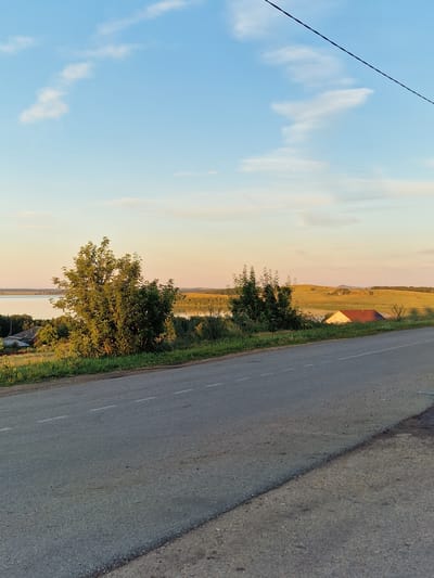 This is a photograph of a rural landscape with a road in the foreground. The road is paved and has dashed white lines marking the lanes. To the left of the road, there is a grassy embankment with some trees and vegetation. In the middle ground, there is a body of water, likely a lake or river, with a small village or settlement on its shore. Beyond the water, rolling hills are visible, with some scattered trees and buildings. The sky is clear with a few wispy clouds, and the overall lighting suggests it is either early morning or late afternoon. The image does not depict any people or specific actions. The location is not the city of Kashgar City, China.