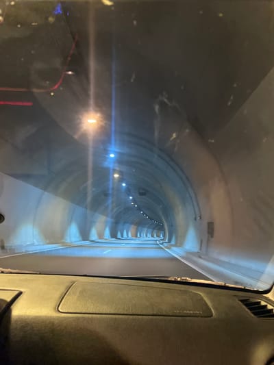The image shows the interior of a vehicle driving through a tunnel. The perspective is from the driver's seat, looking forward through the windshield. The tunnel appears to be well-lit with overhead lights and lights along the tunnel walls. The road surface is visible, with white lane markings. The interior of the car, including the dashboard, is also visible in the foreground. The scene suggests movement through an enclosed passage, typical of a road tunnel. There are no people or specific landmarks visible that would indicate a location in Blatino, Bulgaria.