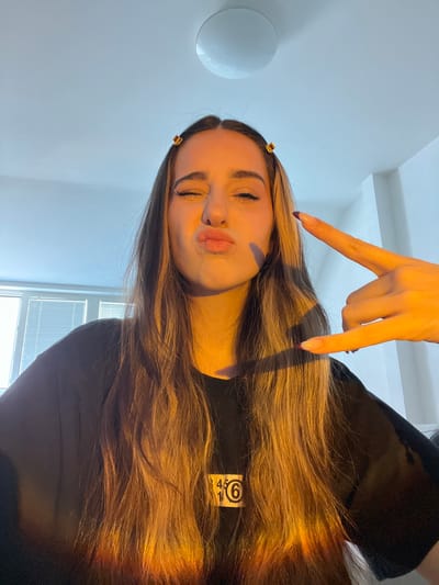 A young woman with long, brown hair, adorned with two small gold hair clips, poses for a selfie in what appears to be an indoor setting. She is making a kissing face and her eyes are closed. Her right hand is raised, forming a "rock on" gesture with her index and pinky fingers extended, while her left hand is not visible in the frame. She is wearing a black t-shirt with a white rectangular logo containing the numbers "45 10 6" and a circle with the number "6" inside. The lighting in the image suggests it was taken during golden hour, with warm light illuminating her hair and casting shadows. In the background, a window with blinds is visible, along with a white ceiling and part of a white wall. The location is identified as the city of Krems an der Donau, Austria.
