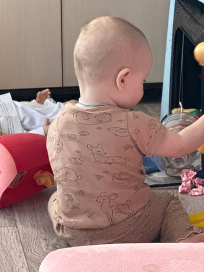A baby lies on a floor next to a large, soft, pink stuffed animal, on a multi-colored, patterned surface. The baby is wearing a light brown shirt with cartoon animal designs and matching pants. The location is not indicated in the image.