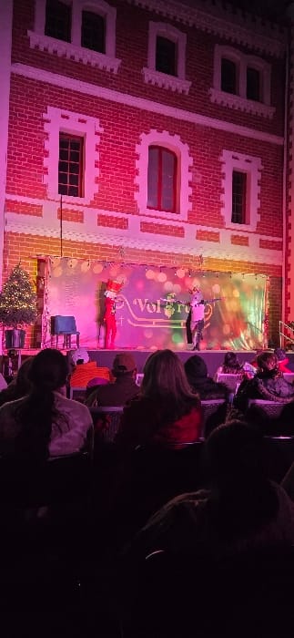 A Christmas show featuring Santa and an elf suspended in the air in front of a building façade backdrop. The building is illuminated with pink light, highlighting brickwork and window details. A banner behind the performers displays the word "Volare". A small Christmas tree sits on the stage below. Audience members can be seen in silhouette in the foreground, watching the performance.