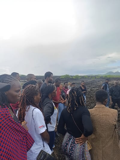 A group of fourteen people are gathered together on a volcanic rock field in Tsavo North National Park, Kenya, located at latitude -2.917 and longitude 38.005. The group is comprised of eight women and six men. They are standing in a semi-circle, facing inward towards the center of the circle. Many are looking at something off-camera, but there is no direct interaction between the group members. The rock field is covered in black volcanic rocks of various sizes, with a few scattered green bushes visible in the background. The sky is overcast with a light grey color, suggesting a partly cloudy day.