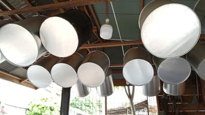 In Geulanggang Teungoh, Indonesia, a low-angle shot captures an interior scene of a structure with a corrugated metal roof supported by wooden beams. Numerous large, cylindrical metal containers, possibly buckets or drums, are suspended from the beams. Most of these containers are silver, reflecting light, and some appear to have white lids or bottoms. One larger, dark-colored container is also visible. A white, bulbous light fixture hangs from the ceiling by a coiled red cord, illuminating the space. The background shows glimpses of outdoor greenery through fencing and the open sides of the structure.