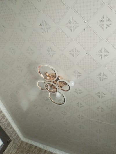 The image shows a close-up, upward-angle view of a modern chandelier hanging from a white textured ceiling. The chandelier features multiple interlocking circular frames with a gold finish, surrounding what appear to be light bulbs. The ceiling has a repeating geometric pattern of diamonds with decorative star-like motifs inside. The lighting is soft and diffused, suggesting an indoor setting. The overall impression is one of contemporary interior decor. There is no visible text in the image.