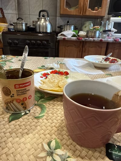A close-up, high-angle shot captures a kitchen table setting in Valday, Russia, featuring a meal and beverages. The table is covered with a woven mat adorned with a floral pattern. In the foreground, a pink mug with a textured design holds dark tea with a tea bag tag that reads "Greenfield." To the left of the mug, an open can of condensed milk, labeled in Russian as "Magnit Varenoe Sgushchennoe Moloko," is visible with a spoon inside. Adjacent to the can is a plate with thin, golden crisps. In the background, a stove with pots and kettles is partially visible, suggesting a domestic kitchen environment. The overall scene depicts a casual moment of consumption.