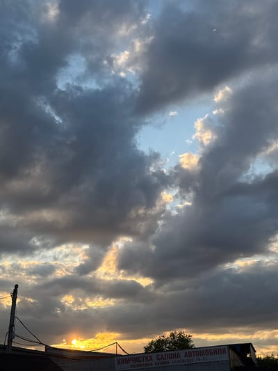 This is an eye-level, outdoor, medium shot of a dramatic cloudy sky at sunset, taken in Sovkhoza "Ramenskoe", Russia. The sun is setting behind a building with a visible sign that reads "ХИМЧИСТКА САЛОНА АВТОМОБИЛЯ ПОЛИРОВКА КУЗОВА" which translates to "Car Interior Cleaning, Body Polishing". A utility pole with wires is visible on the left. The clouds are dark and stormy, with patches of sunlight breaking through, creating an orange and yellow glow. The overall mood is ominous yet beautiful. There are no people or animals visible in the image.