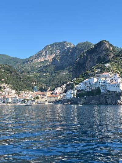A scenic view of the Amalfi Coast in Pogerola, Italy, showcases the picturesque town nestled against lush, mountainous terrain. White buildings with red-tiled roofs cascade down the steep slopes towards the azure sea. The water in the foreground is a deep blue, rippling with sunlight, reflecting the vibrant sky. A sailboat with dark sails is anchored in the bay, and a small white boat is visible near the shore. The backdrop is dominated by rugged mountains covered in dense green vegetation, with terraced slopes suggesting agricultural activity. The sky above is a clear, bright blue, indicating a sunny day.