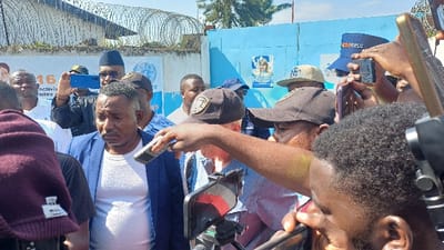 In Kabinzi, Rwanda, a dense outdoor gathering focuses on a somber-faced man of medium complexion and dark hair, wearing a blue blazer over a white t-shirt, positioned centrally. He is surrounded by numerous individuals, predominantly male, many actively holding smartphones vertically to record or photograph the scene. One hand extends a rectangular silver and black microphone towards the central figure. A smartphone mounted on a stand is visible in the immediate foreground. Among the onlookers, several wear distinct headwear: a dark baseball cap with a gold emblem, a dark blue cap with "BASSWORLD" text, and a dark blue police cap with "POLICE" visible. The background reveals a light-colored concrete wall topped with coiled razor wire. Below the razor wire, a white banner is partially visible, displaying "16," "activités," and a United Nations logo. Adjacent to this, a vibrant blue corrugated metal wall or gate features a poster of a woman with blonde hair. The scene is illuminated by natural daylight under a clear sky.