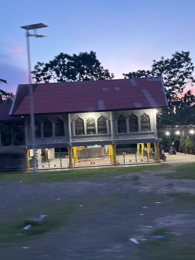 The image depicts a stilted building with a red tiled roof, reminiscent of a mosque or community hall, situated in Matangpanyang, Indonesia. The building is elevated on yellow and gray pillars and features multiple arched windows with intricate metalwork. The sky above displays a gradient of purple and pink hues, indicating twilight. A solar-powered street lamp stands to the left of the building. In the foreground, a grassy area is visible with some scattered debris. A black fence encloses the area around the building. To the right, a paved area shows two motorcycles and several outdoor lights. The overall scene is tranquil, capturing a moment at dusk in a rural or semi-rural Indonesian setting.