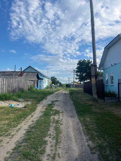 This is a vertical, eye-level, daytime photograph taken in Сухринское, Russia. The image captures a rural residential street scene. A dirt road runs diagonally from the bottom right to the top center of the frame, flanked by grassy areas and houses.

On the left side of the road, in the foreground, is a weathered, wooden house with a corrugated roof. A metal pipe protrudes from the roof. Further down the street, another house painted light blue with white trim is visible, featuring several windows. A dark fence separates this property from the road. A large wooden utility pole with wires stretches across the upper left portion of the image.

On the right side of the road, the grassy embankment is interspersed with patches of bare dirt. Further down the street, more houses and trees can be seen. In the far distance, the road continues into a cluster of trees. There are some plastic bottles and children's toys scattered along the edge of the road on the right.

The sky is bright blue with scattered white, puffy clouds, suggesting a clear and sunny day. The lighting indicates it is likely midday. The overall mood is tranquil and everyday, reflecting a quiet village life. No people are visible in the image. There is no visible text.