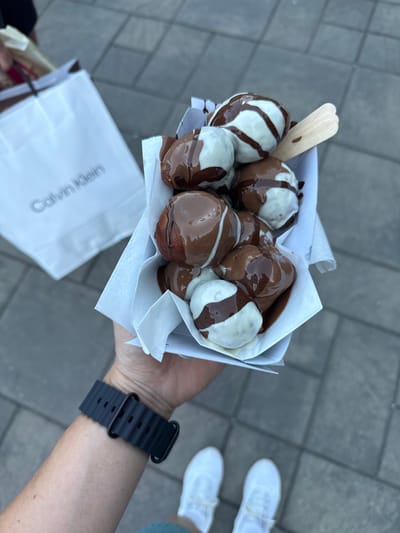 A person is holding a white paper container filled with dessert items that appear to be dough balls coated in chocolate and white sauce. The dessert is presented in a white paper holder, with two wooden popsicle sticks inserted into the container. In the background, a white shopping bag with "Calvin Klein" logo is visible, placed on a gray paved surface. The scene is captured from a first-person perspective, with the person's wrist, wearing a black watch, and their white sneakers visible at the bottom of the frame. The location is not identifiable as Verpet, Norway.