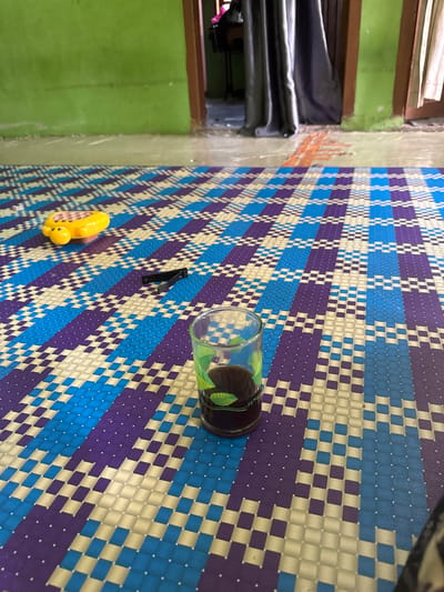 In Babahjurong, Indonesia, an indoor scene is depicted with a focus on a woven mat on the floor. The mat features a repeating geometric pattern of blue, purple, and off-white squares. On the mat, a small yellow toy, possibly a duck, and a pair of black nail clippers are visible. Towards the foreground, a clear glass contains a dark liquid, possibly coffee or tea, and a green leaf decoration is visible inside the glass. The background shows a green wall and a doorway leading to another room, with curtains drawn. The lighting suggests it is daytime.