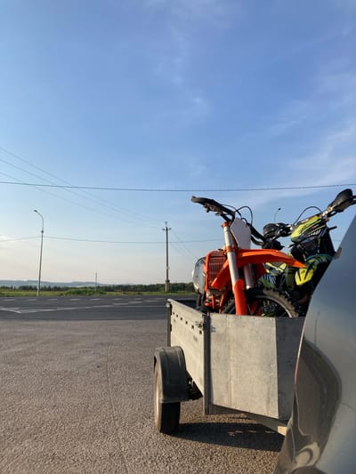 The image shows two motorcycles on a trailer, parked on an asphalt road. The road appears to be a highway or major thoroughfare, with road markings visible. Power lines and poles run alongside the road. In the background, there's a glimpse of vegetation, possibly trees and hills, suggesting a rural or semi-rural setting. The sky is blue with some wisps of clouds. The location is Verkh-Sosnova, Russia.