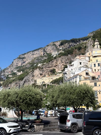 The image depicts a street scene in Pogerola, Italy, set against a backdrop of a steep, rocky cliff face dotted with buildings. The sky above is clear and blue.

A row of parked cars lines the right side of the street, including a dark van and a white SUV with its rear door open. Two large, bushy trees stand between the cars and the cliff face, partially obscuring the view of the buildings.

On the street, a partially visible water feature or fountain stands next to a parked scooter. People are walking around, suggesting it is an active public space. The buildings visible on the cliff are a mix of white and yellow structures.