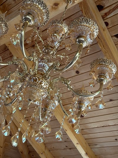The image shows a complex, multi-tiered chandelier hanging from a wooden ceiling in Gornje Mrke, Montenegro. The chandelier is made of ornate metal fixtures and cut glass or crystal pendants. The ceiling is constructed of light-colored wooden planks and beams. The lighting suggests an indoor environment.