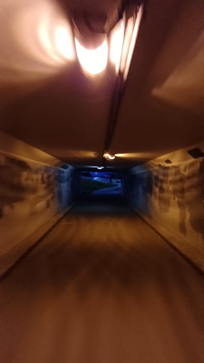 The image shows a narrow, dimly lit corridor or tunnel. Walls are brown and appear to be concrete. Light sources on the left wall cast bright, warm light. The far end of the corridor is darker with blue and green light, and possibly a curtain or other fabric. The photograph appears to be taken from a low angle with a sense of motion, as if the camera is moving forward through the corridor. There is no indication in the image that suggests the location is Obukhovo, Russia. There are no visible people or identifiable subjects.