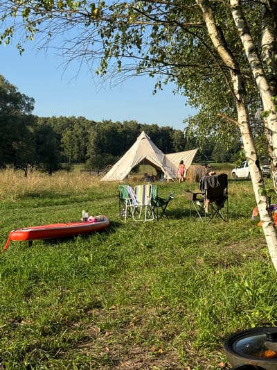 This image depicts an outdoor scene in Kuzemkino, Russia, likely a campsite. In the foreground, a red inflatable paddleboard rests on green grass. Several camping chairs are also present, some unoccupied and others with people seated. A large, off-white bell tent is visible in the middle ground, with figures near its entrance. Behind the tent, a round hay bale is situated, and a white car is parked to the right. A line of trees forms the background, with a clear blue sky above. The overall environment is grassy and appears to be a rural or park-like setting.