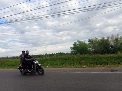The image depicts two individuals riding a scooter on a road in what appears to be a rural area near Sagoe, Indonesia. The road surface is asphalt and has a white line marking the lane. A grassy verge separates the road from a field of vegetation, with trees and shrubs visible in the background. Overhead, power lines stretch across the sky, which is filled with clouds. The foreground of the road shows some dark patches. The scooter and its occupants are in motion.