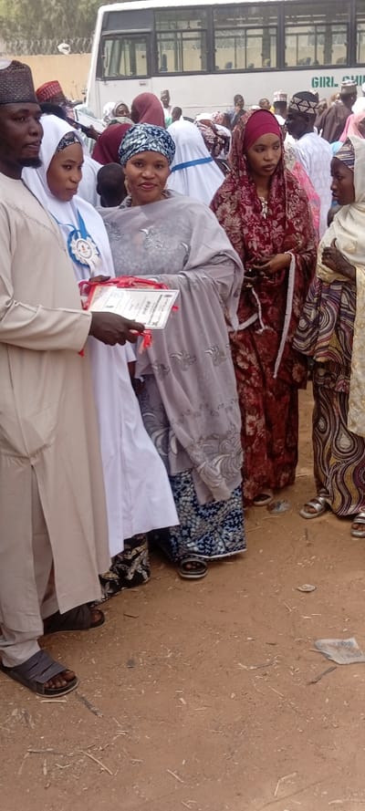The image captures an outdoor gathering in Sani Mai Nagge, Nigeria, centered around a small group of individuals on a light brown dirt ground. In the foreground left, a man in a light-colored caftan and dark cap holds a white certificate with red ribbons in his left hand. To his immediate right, a woman in a white hijab and dress wears a blue and white decorative ribbon with a medallion around her neck, looking towards the certificate. Adjacent to her, a woman in a grey patterned headscarf and top, worn over a blue patterned skirt, smiles directly at the viewer, her right hand positioned near the certificate. Further right, a woman in a dark red patterned hijab and dress stands, her gaze directed to the right. On the far right, another woman wears a light beige head covering and a patterned dress, displaying visible henna designs on her hands and wearing silver sandals, looking towards the left. The ground is uneven, littered with small sticks and debris. In the background, a white bus with the words "GIRL CHI" (likely "GIRL CHILD") partially visible in green lettering is parked, behind which a crowd of indistinct individuals and structures are present under a bright sky.