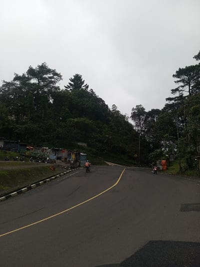 The image depicts a two-lane asphalt road under an overcast sky in Deothang, Bhutan. The road, marked by a continuous double yellow line down its center, curves gently to the right and left, forming a split or intersection ahead. A black and white striped curb borders the left edge of the road, alongside a grass-covered embankment.
To the immediate left of the road, behind the embankment, a series of rudimentary market stalls or shelters constructed from corrugated metal, tarpaulins, and various other materials are visible. Multiple motorcycles are parked adjacent to these structures. A dense, verdant hillside covered with trees, including prominent pine-like evergreens, rises sharply behind the stalls and dominates the left background.
On the road, a person wearing a light-colored helmet and shirt rides a light-colored motorcycle away from the viewer on the left side of the road. Further down the road, another person on a dark motorcycle with red accents is visible on the right side, approaching the viewer. The right side of the road is lined with dense green trees and foliage, with a utility pole visible amongst them. Some orange tarp-covered objects are present near the trees on the right further down the road. The sky is uniformly light grey, indicating overcast conditions.
