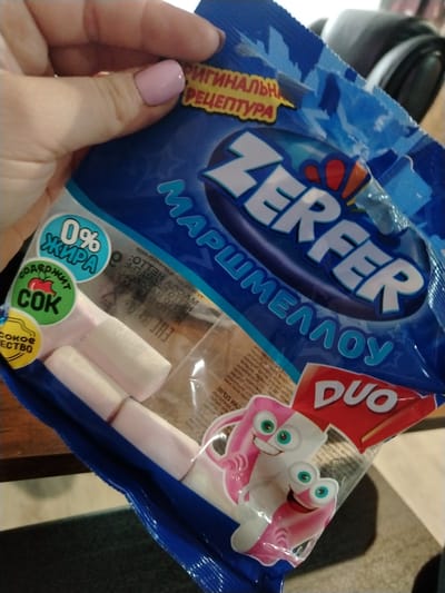 A hand, adorned with light pink nail polish, holds a blue and white package of "ZERFER" marshmallows. The package features the brand name "ZERFER" prominently in white letters with red and blue accents, along with "МАРШМЕЛЛОУ" (Marshmallow) and "DUO" in a red triangle at the bottom right.
The packaging highlights several features: a yellow banner partially visible at the top reads "ОРИГИНАЛЬНАЯ РЕЦЕПТУРА" (Original Recipe). Two circular badges on the left side indicate "0% ЖИРА" (0% FAT) in blue and "СОДЕРЖИТ СОК" (CONTAINS JUICE) with an apple icon in green. Below the green badge, another partially visible yellow badge says "СОКОВОЕ ВЕЩЕСТВО" (Juice substance).
Through a clear window in the packaging, several pink and white cylindrical marshmallows are visible. The bottom right of the package displays cheerful cartoon marshmallow characters, one white and one pink, with large blue eyes. The background shows a dark wooden surface, possibly a table, and a darker, textured surface below, likely a floor or rug. The image is taken indoors, showing the product in a casual setting. The text on the packaging is in Russian, consistent with the provided location context of Yaroslavl', Russia.