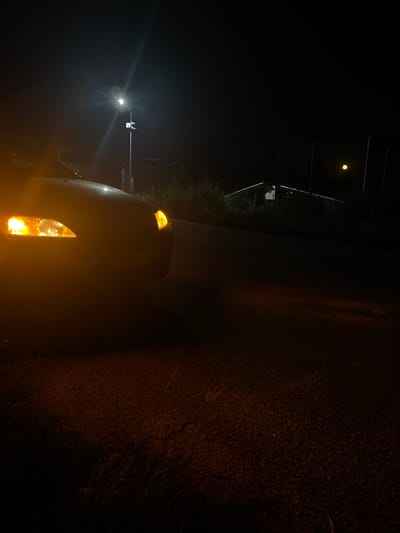 The image captures a nighttime scene on a road, likely in Vedrare, Bulgaria. The primary subject is a vehicle, possibly a van or truck, with its hazard lights and headlights illuminated. The warm glow of the lights cuts through the darkness, casting reflections on the vehicle's surface and illuminating the road ahead and to the side.
The road appears to be paved, with the right side bordered by an unpaved area covered in gravel and vegetation, suggesting a rural or semi-rural setting. In the distance, a streetlamp or overhead light is visible, casting a bright glare and contributing to the overall low-light atmosphere. Above, the faint outline of trees can be seen against the dark sky. A small, luminous object, possibly the moon, is visible in the upper left portion of the frame.
There are no people visible in the image. The activity taking place is implied to be driving or stationary on the side of the road at night. Notable details include the time of day, which is clearly night, and the potential for a clear or mildly overcast sky given the visibility of the moon. The setting appears to be a road in a somewhat undeveloped area, judging by the unpaved shoulder and vegetation. The emotional tone is one of quiet darkness punctuated by artificial light.
There is faint, illegible text visible on the side of the vehicle, appearing as a white or light-colored inscription against the darker paint.