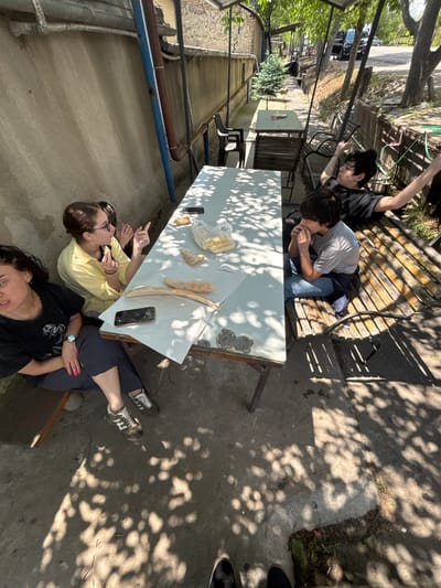 In Badiauri, Georgia, four people are seated around a white table outdoors. A woman in a yellow shirt and glasses is holding food and speaking, gesturing with her other hand. Next to her, a woman in a black t-shirt is looking towards the camera. On the other side of the table, two young males are seated. One, in a grey t-shirt and jeans, is eating. The other, in a black t-shirt, is reclining on a wooden bench with his arms outstretched. The table is covered with food, including bread and a plastic bag of snacks, and a mobile phone. The scene takes place in a shaded area with trees casting dappled sunlight on the ground. A wall and a fence are visible in the background, along with a road and parked cars. The ground is concrete.