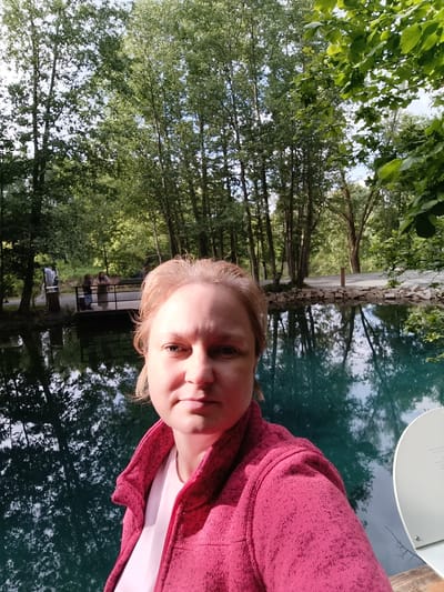 This is a selfie taken outdoors by a woman with blonde hair. She is wearing a pink jacket over a white shirt. The background shows a tranquil scene with a body of clear blue water, likely a lake or pond, reflecting the surrounding trees. On the far side of the water, there is a wooden deck or pier with a few people standing on it, one of whom appears to be a child. The area is densely wooded with tall trees, suggesting a natural park or a forest setting. The lighting indicates it is daytime, likely late morning or afternoon, with soft sunlight filtering through the leaves. The overall mood of the image is peaceful and serene. There is no visible text in the media file.