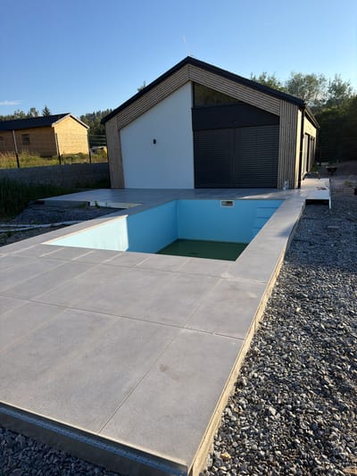 The image shows an outdoor scene in Horní Planá, Czechia, on a sunny day. In the foreground, a grey tiled patio surrounds an empty swimming pool with blue interior walls. The pool appears to be under construction or renovation, with a small section of green water visible at the bottom. Beyond the patio, a modern house with white and wooden facade stands, featuring large dark-colored shutters. In the background, another smaller building with a wooden facade and a dark roof is visible, along with trees and a clear blue sky. The ground around the patio and house is covered in grey gravel. There are no people or animals visible in the image.