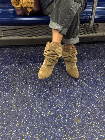 The image shows a person seated on what appears to be public transportation, possibly a subway or train. The person is wearing cuffed gray jeans and slouchy tan boots. Their feet are extended onto the speckled blue and yellow floor of the vehicle. A metal safety bar and a perforated metal panel are visible to the left of the person. A brown knitted item rests on the seat.
Since the prompt includes the city of Hoboken, United States of America, the image could be from the Port Authority Trans-Hudson (PATH) train that serves the city. However, this cannot be confirmed from the image alone.