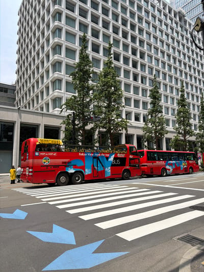In Chiyoda, Japan, a scene unfolds on a wide street with a prominent, modern building featuring a grid-like facade dominating the background. In the foreground, two red, open-top tour buses are parked side-by-side. The bus on the left is a double-decker, with "SKY BUS" and "HOP on HOP off" branding visible. Its side displays "Tokyo View from the top" in large blue and white lettering against a sky and cloud graphic. The bus on the right, also red, has "HOP on HOP off" and "SKY BUS" lettering.

A pedestrian crosswalk, marked with bold white stripes, spans the street in front of the buses. On the road surface, several large blue directional arrows point towards the crosswalk.

To the left of the buses, two individuals are visible on the sidewalk. One person, wearing a bright yellow coat, stands facing away from the camera. Beside them, another person, wearing a white shirt and dark pants, is also present.

The environment suggests a bright, possibly daytime setting, with clear skies overhead. The architectural style of the building and the presence of tour buses indicate a tourist-oriented area within the city.