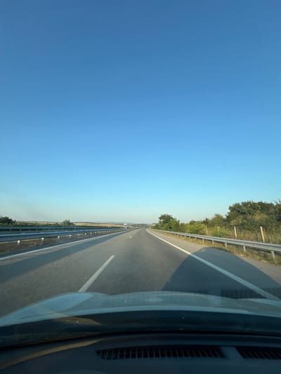The image depicts a view from inside a vehicle traveling on a highway. The highway stretches into the distance, with a clear blue sky above. Guardrails line both sides of the road, and vegetation can be seen beyond them. In the distance, a truck is visible on the highway. The dashboard and windshield of the vehicle are visible in the foreground. The scene appears to be set in the countryside, possibly near the city of Aleksandrovo, Bulgaria, given the context. There are no people or animals in the image. The lighting suggests it is either early morning or late afternoon.