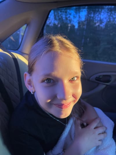 A young Caucasian girl with blonde hair is sitting in the back seat of a car, looking at the camera and smiling. She is wearing a black shirt and a silver necklace. The background shows the interior of the car and a blurred view of trees outside the window. The lighting suggests it is either dusk or dawn, with a purple and blue hue cast over the scene. The location is Makarovka, Russia.