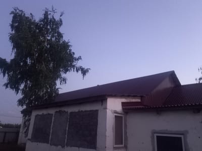 A medium shot captures a modest, white, stucco building with a dark red corrugated metal roof under a pale, hazy twilight sky. The building's facade is partially covered with dark, textured patches, possibly indicating repair or unfinished work. Two boarded-up windows are visible on the left side of the building, while a single, smaller window with a light-colored frame and dark pane is present on the right. A large, leafy tree with dark foliage leans over the left side of the building, its branches extending towards the roof. In the background, faint silhouettes of other structures and trees are visible on the horizon, suggesting a rural or semi-rural setting. The overall impression is one of a utilitarian structure in a quiet, dimly lit environment.