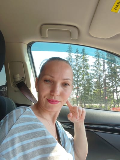 A woman is sitting in the driver's seat of a car. She is wearing a striped gray and light blue shirt and has a gold necklace. She is looking directly at the camera with a hand raised near her cheek. Tall pine trees are visible through the car window. The car is located in Пажга, Russia.
