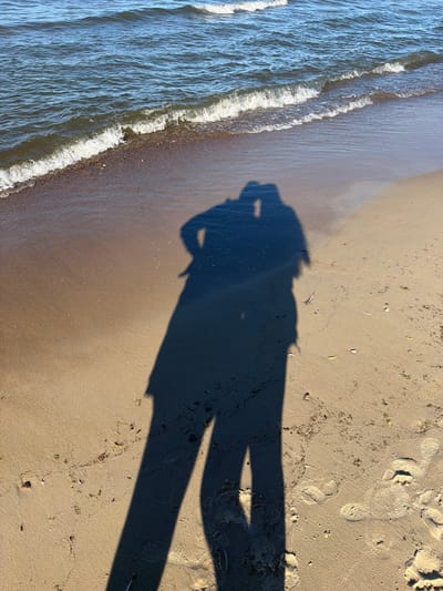 The image shows two shadows of people on a sandy beach next to the sea. The sea has gentle waves with white foam. The sand is wet near the water and drier further up the beach, with some small pebbles and debris scattered on it. Footprints are visible in the sand. The shadows suggest the people are standing close together, possibly in an embrace or looking at each other. The lighting indicates it is daytime, with the sun casting long shadows. The scene is peaceful and evokes a sense of a shared moment by the water. The location is identified as the beach in Zīvartiņš, Latvia.