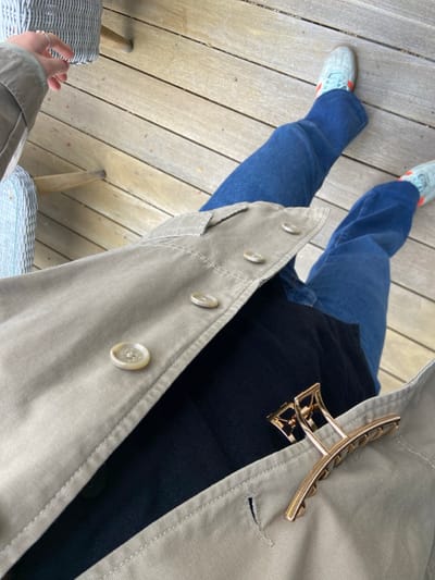 The image shows a person's lower body, wearing a grey jacket and blue jeans, and a black shirt underneath the jacket, standing on a wooden deck. The person is wearing a pair of blue sneakers with orange accents. A gold-colored hair clip is attached to the jacket. The person is wearing a ring on their left hand. The location of the image is Rye, New York, United States. The wooden deck is slightly dirty and worn.